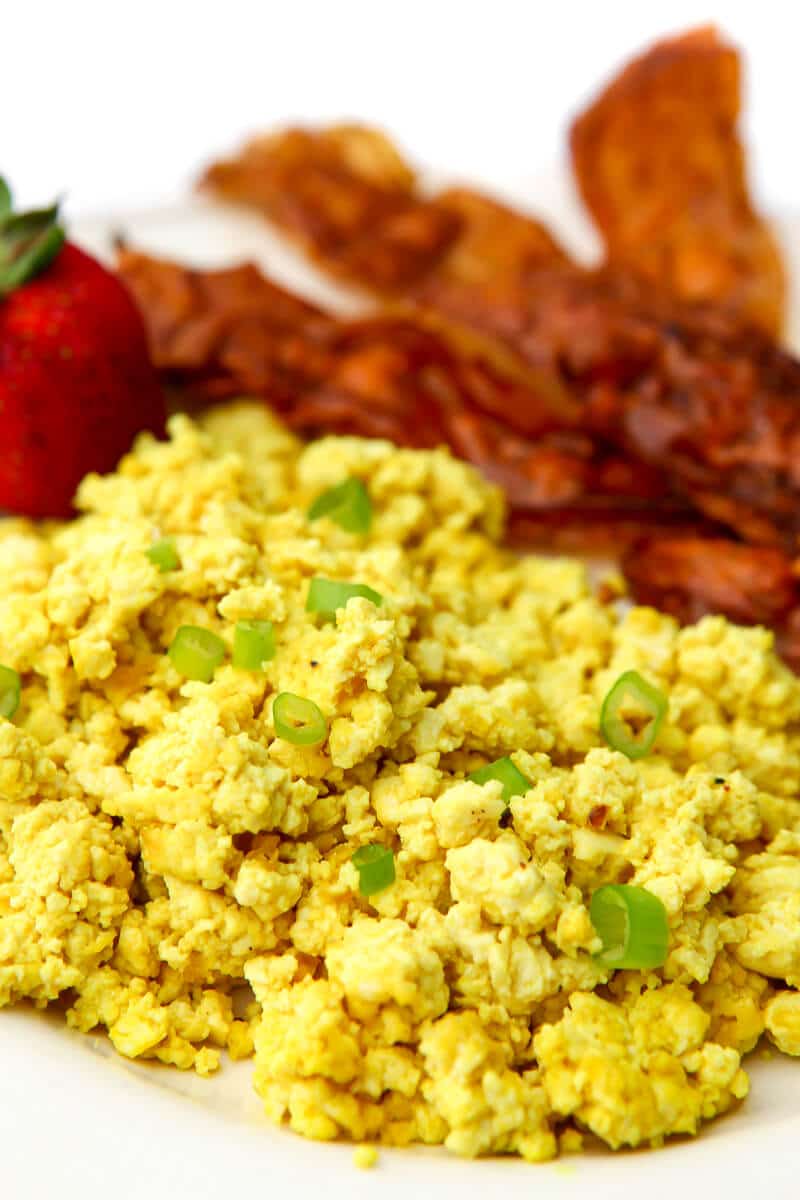 Huevos revueltos veganos hechos de tofu cubiertos con cebollas verdes con tocino vegano y una fresa al lado.